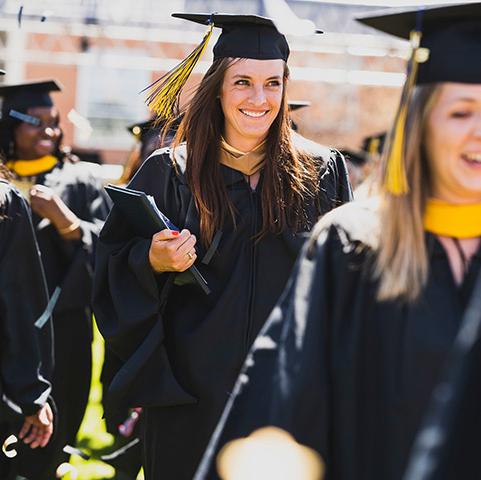 a master's graduate smiles while wearing regalia and recessing from the stage with her peers at the Spring commencement ceremony on the Quad on the Northwest Denver campus