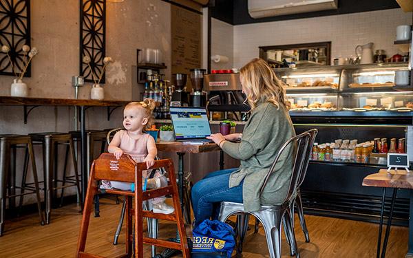 一位正在用笔记本电脑工作的妇女坐在咖啡店桌旁的高脚椅上，旁边是一个蹒跚学步的孩子
